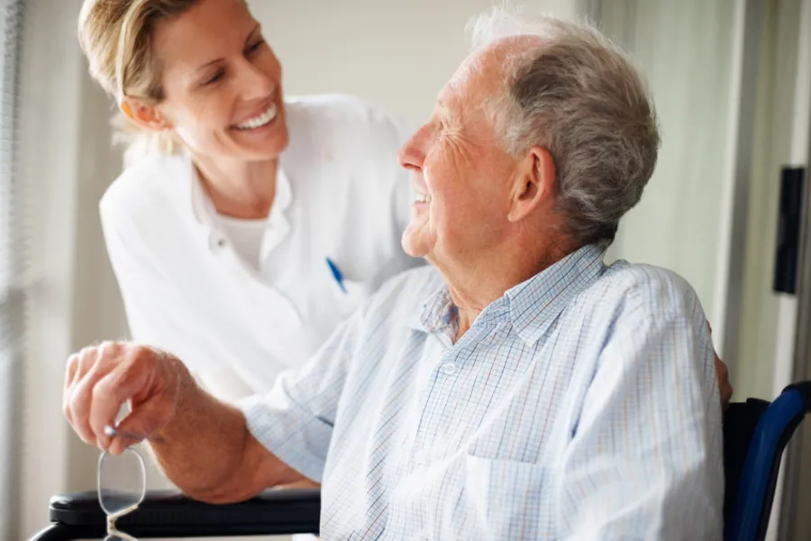 Eldre mannlig pasient i rullestol og smilende kvinnelig ansatt i hvit frakk som kommuniserer