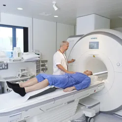 En pasient ligger på benk ved CT maskin med ansatt stående ved siden av