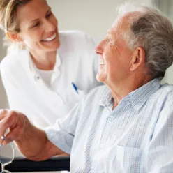 Foto av en gammel mann i stol som ser opp på en smilende kliniker i hvit frakk.