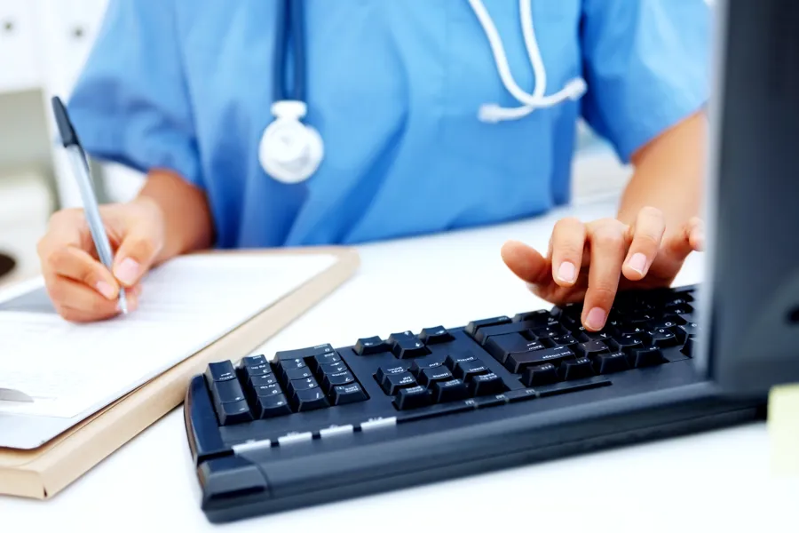 Bilde av ansatt i blå skjorte med stetoskop rundt halsen, tastatur til pc og penn og papir